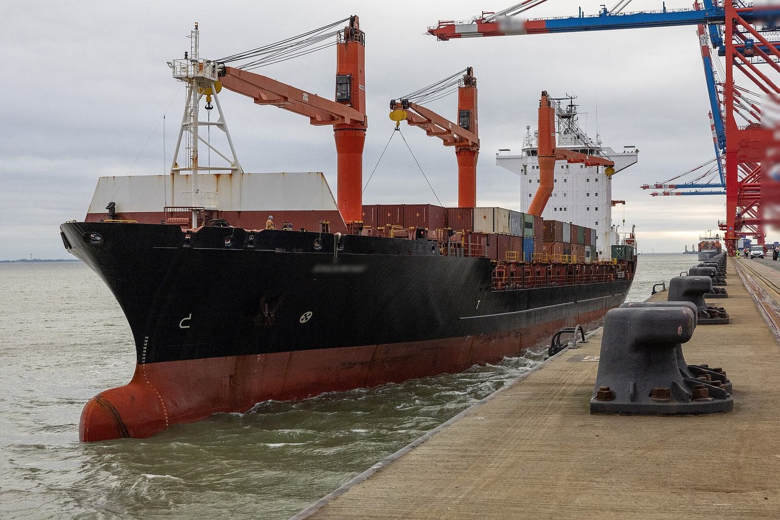 Container ship with deck cranes arriving alongside a quay beneath port gantry cranes